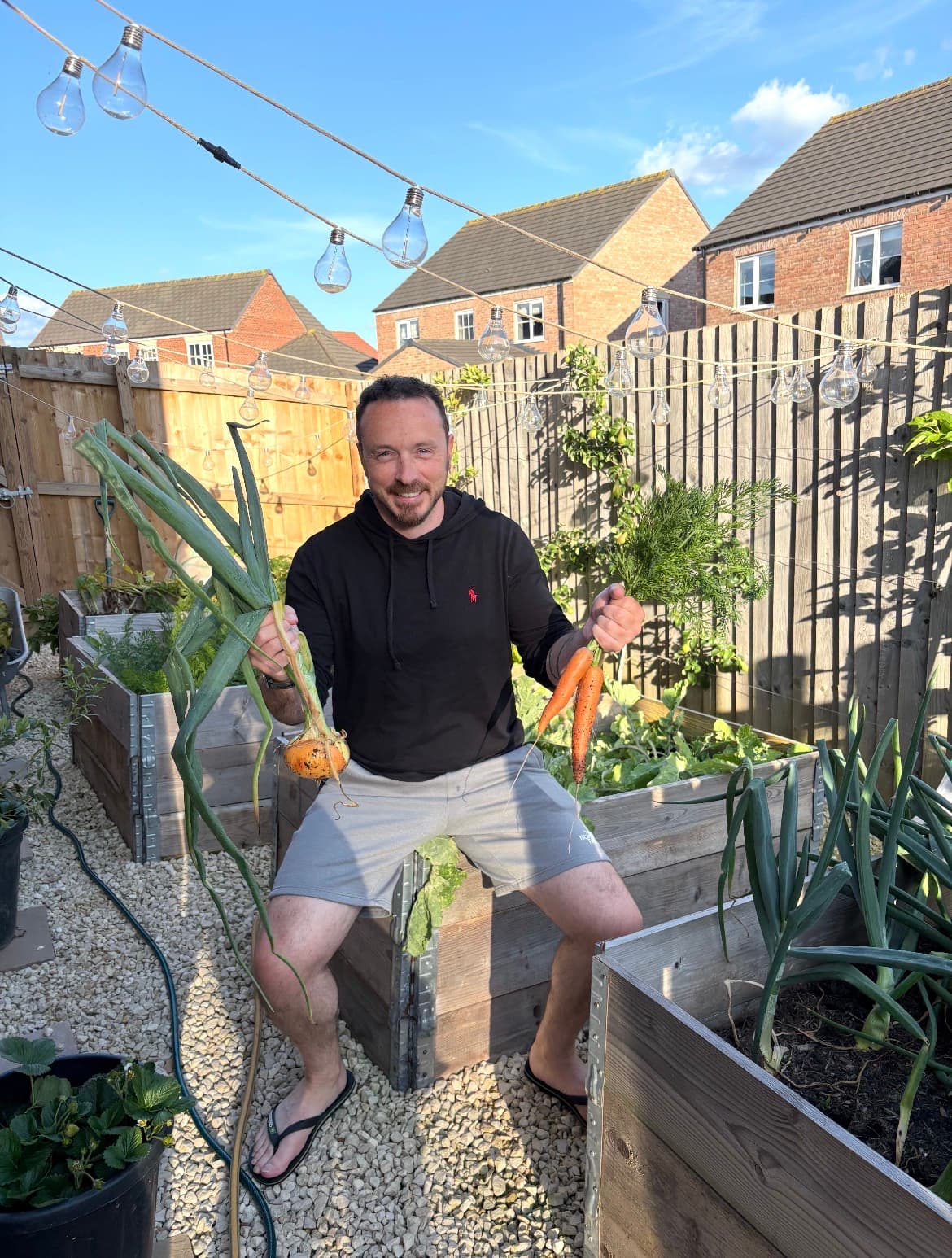 Mark, Vercro’s founder, sitting on a timber raised bed in his back garden holding an onion and a bunch of carrots.